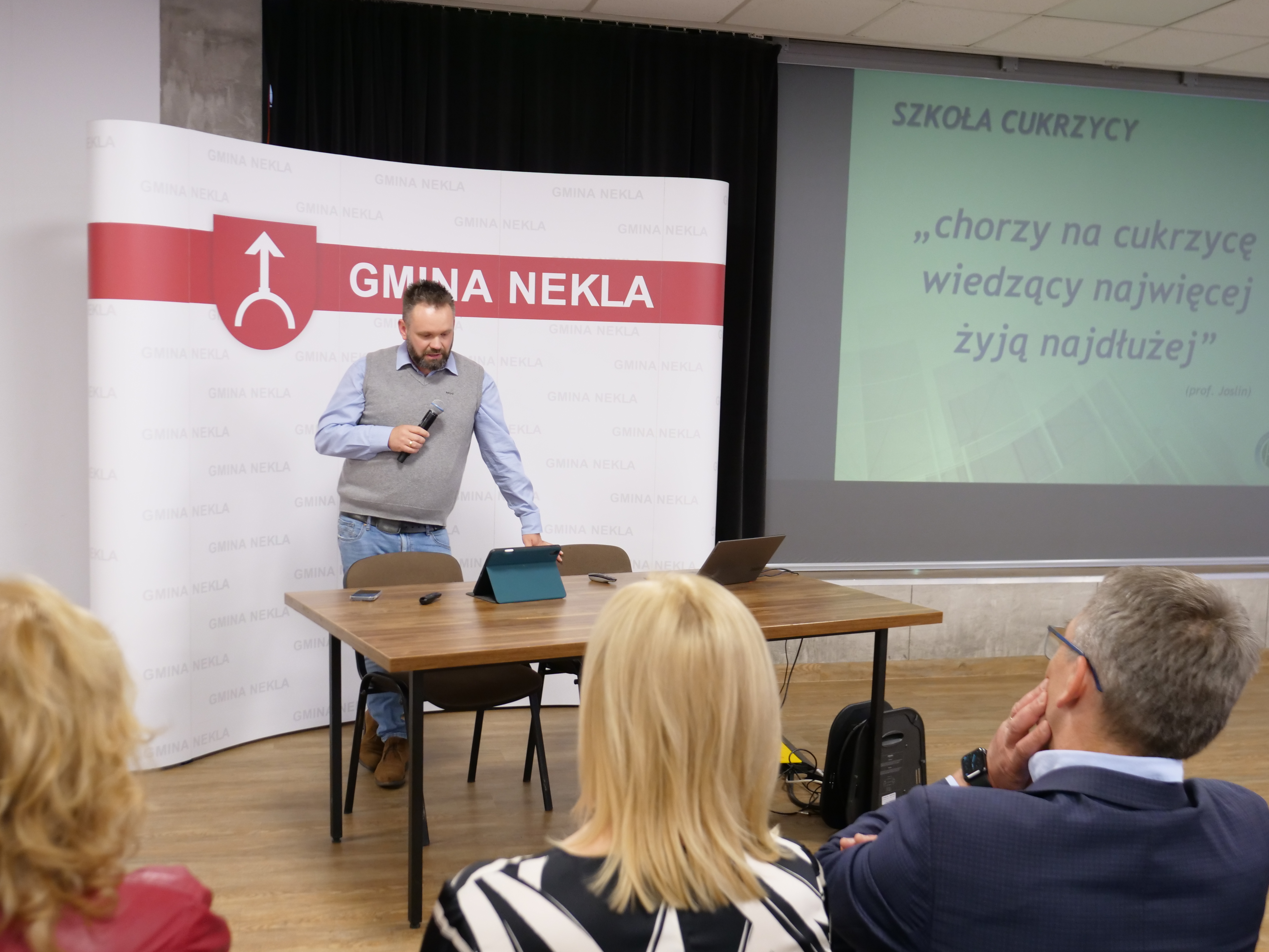 Mężczyzna z mikrofonem w ręku przemawia do grupy os&oacute;b. Za nim baner z napisem Gmina Nekla. Obok na rzutniku napis: Szkoła Cukrzycy, 