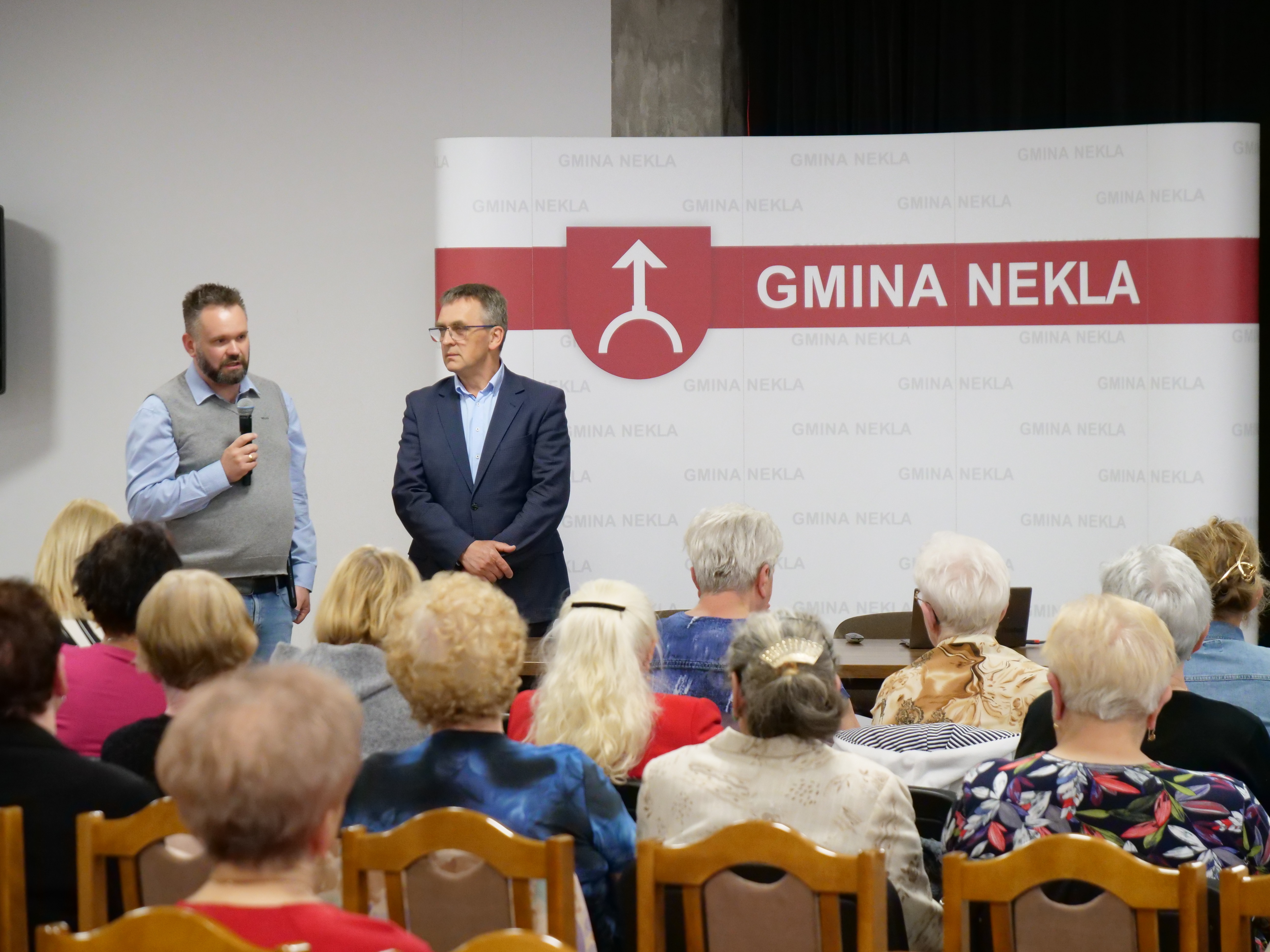 Dw&oacute;ch mężczyzn przemawia do grupy os&oacute;b, siedzącej na krzesłach. Jeden trzyma mikrofon. W tle baner z napisem Gmina Nekla.