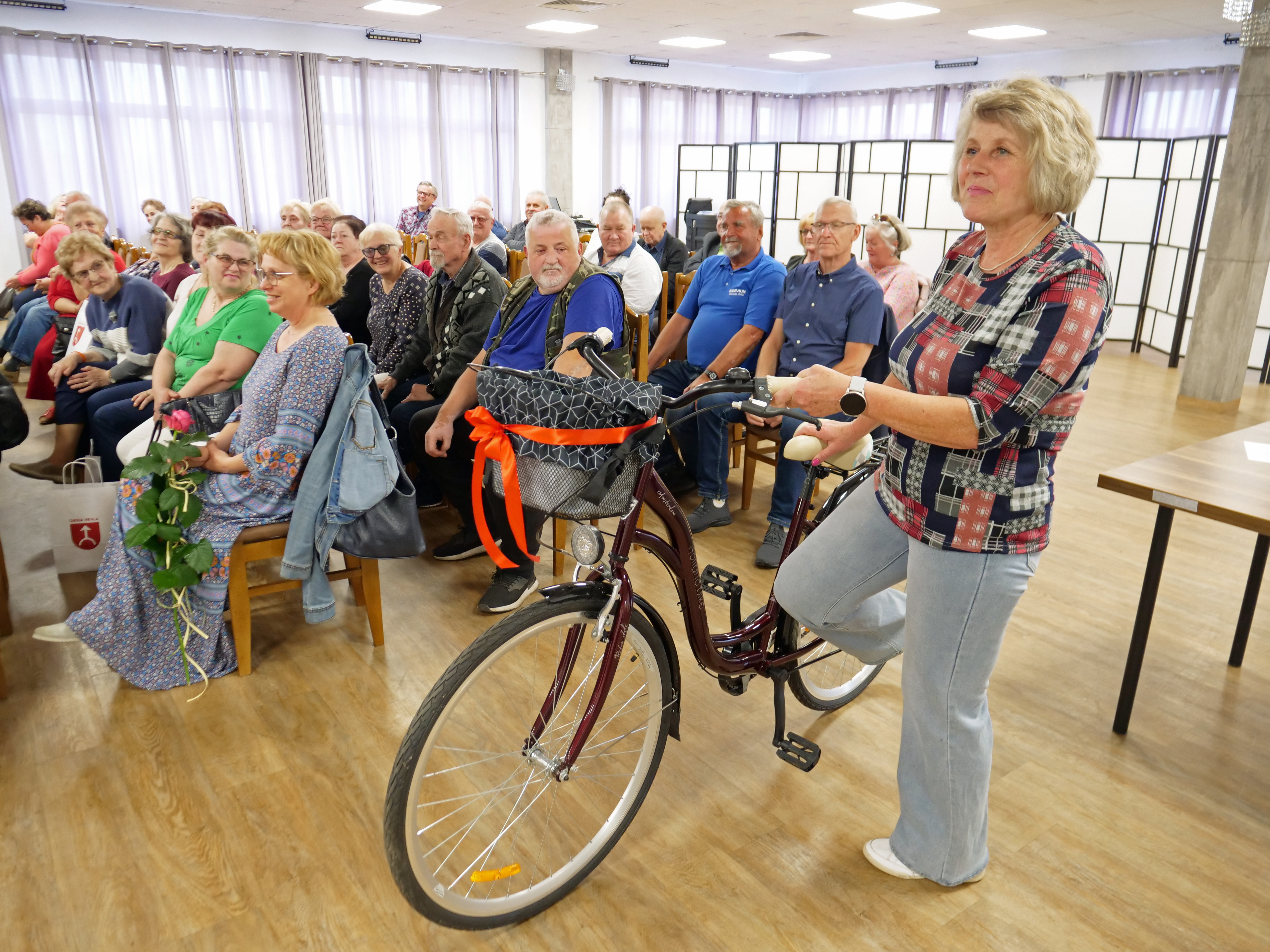 Na pierwszym planie kobieta trzymające kierownicę roweru. W tle grupa os&oacute;b na krzesłach, ok. 30 os&oacute;b.