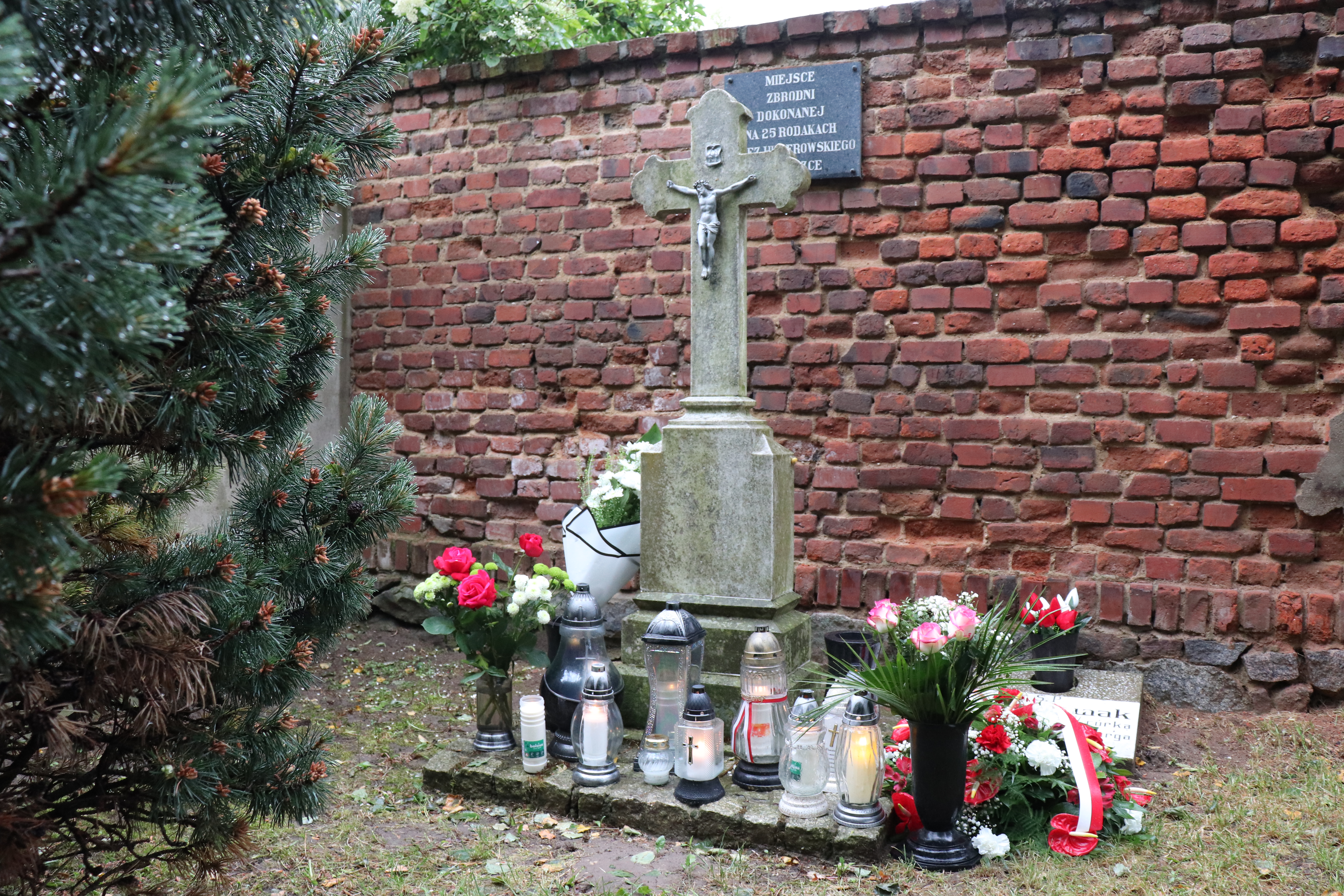Miejsce rozstrzelań. Na pierwszym planie kamienny krzyż, znicze i kwiaty. W tle ściana z czerwonej cegły, przy kt&oacute;rej rozstrzelano Polak&oacute;w. 