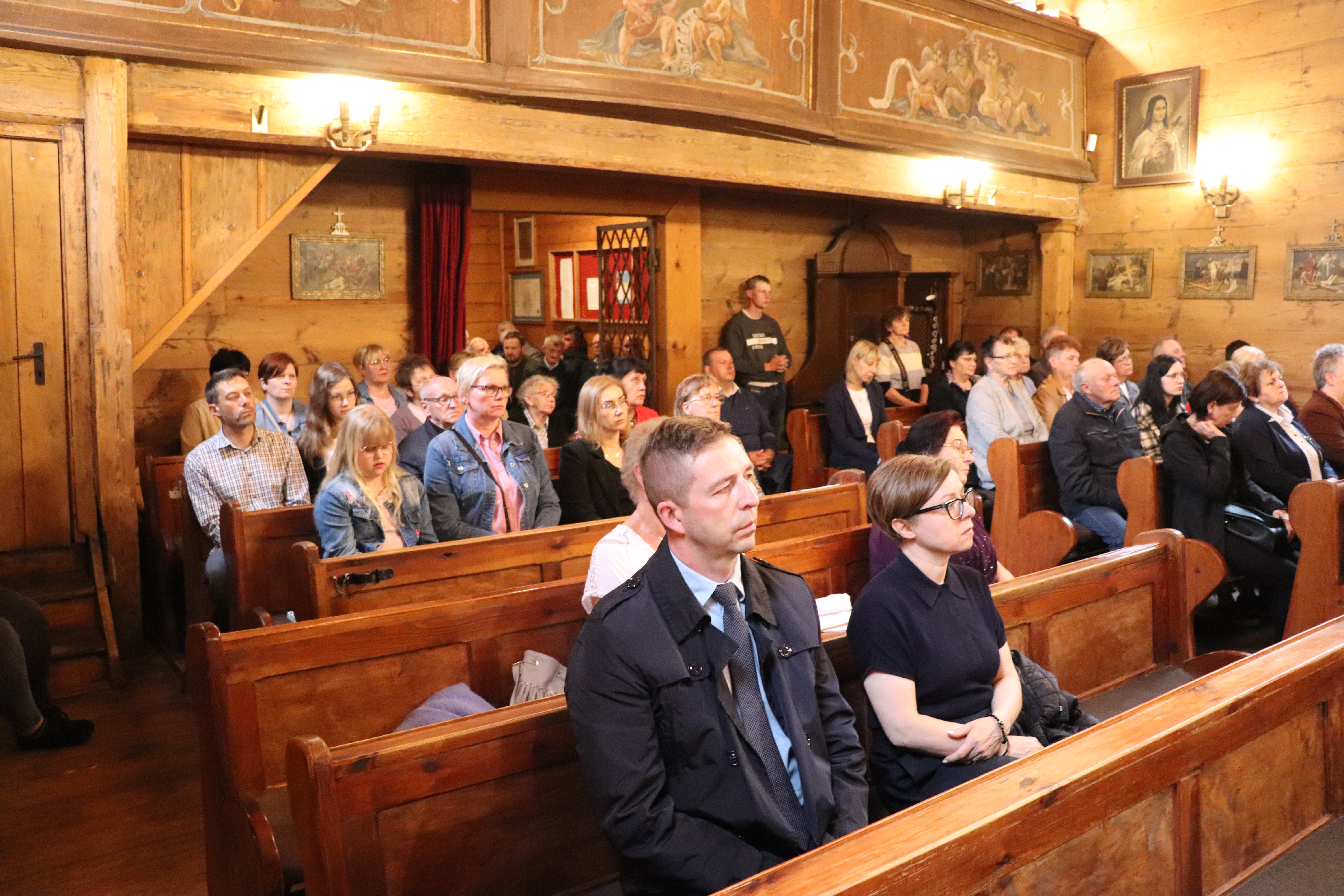 Wnętrze drewnianego kościoła w Opat&oacute;wku. W drewnianych ławkach siedzi kilkadziesiąt os&oacute;b, kobiety i mężczyźni. Wszyscy są zasłuchani.