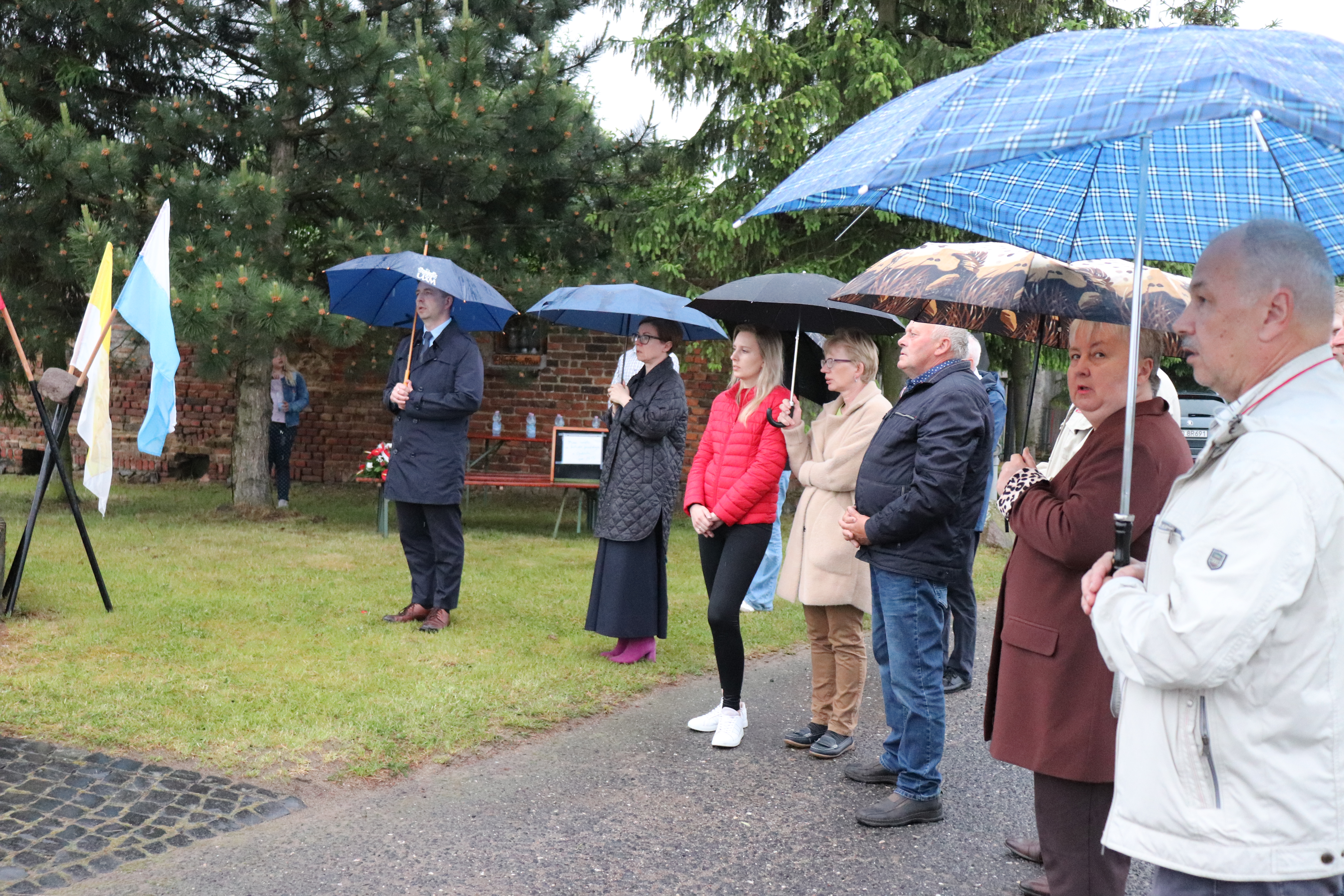 Grupa os&oacute;b w trakcie modlitwy przy pomniku, wszyscy trzymają parasole.
